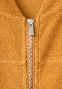 Prim-plan al unui articol de îmbrăcăminte din material portocaliu texturat, cu un fermoar metalic argintiu centrat, parțial închis.