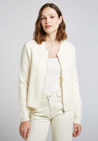 Gilet zippé blanc en tissu texturé, avec des poignets et un col côtelés. Porté sur un haut ajusté blanc et un pantalon de couleur claire.