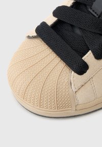 Μπεζ αθλητικό παπούτσι με υφασμένη καουτσούκ μύτη και μαύρα κορδόνια, με σουέντ λεπτομέρειες και σόλα με σχέδιο για πρόσφυση και υποστήριξη.