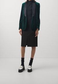 Grön sammetsblazer med svarta satinrevers, kombinerad med en svart knälång kjol och svarta strumpor, samt vita och svarta plattformsskor.