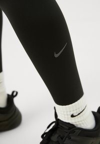 Leggings atletici neri con una superficie liscia e un discreto logo Nike grigio sulla parte inferiore della gamba, abbinati a calzini bianchi a coste e scarpe nere.