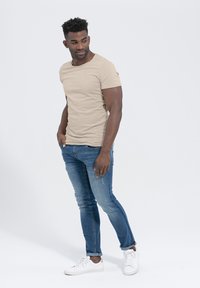 Beżowa dopasowana koszulka z krótkim rękawem, zestawiona z niebieskimi dżinsami slim-fit i białymi sneakersami. Minimalistyczny design, miękka tekstura, casualowy styl.