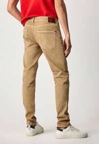 Pantalones de algodón beige con un corte ajustado, que presentan bolsillos traseros, un parche de cuero y detalles de costura. Combinados con zapatillas blancas.