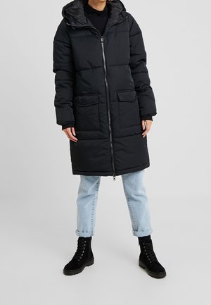 Personne portant un long manteau matelassé noir avec une capuche, un jean bleu clair retroussé et des bottes noires à lacets, debout sur un sol gris.