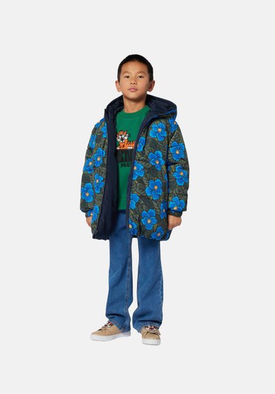 Veste rembourrée avec un motif floral bleu sur fond sombre, avec une capuche. Portée sur une chemise verte et un jean bleu, avec des chaussures beiges.