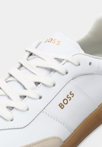 Sneaker alb din piele cu accente bej, având branding auriu "BOSS", șireturi plate albe și o talpă din cauciuc.