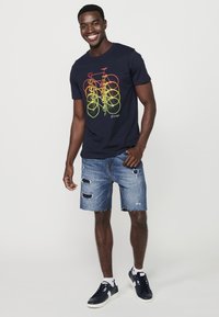 Camiseta azul marino con un gráfico de bicicleta multicolor, combinada con pantalones cortos de denim desgastados y zapatillas oscuras. Diseño sencillo, ajuste casual.