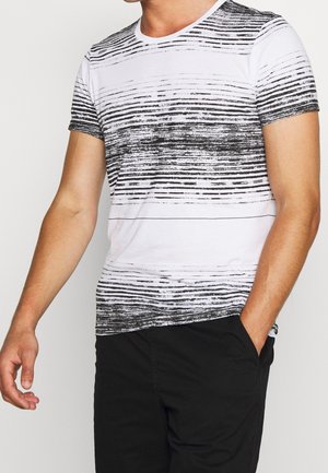 Mand iført hvid kortærmet skjorte med sorte, horisontale teksturerede striber og sorte bukser, med hånden i lommen, foran en ensfarvet baggrund.