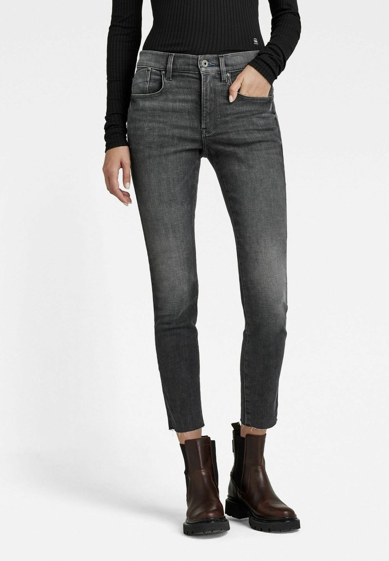 Femme portant un jean noir délavé slim, un haut noir côtelé à manches longues et des bottines marron foncé, la main dans la poche.