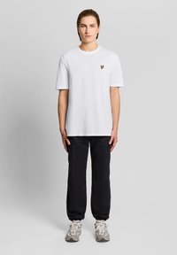 Νεαρός που στέκεται, φοράει ένα απλό λευκό μπλουζάκι με μικρό κίτρινο λογότυπο, μαύρο παντελόνι και γκρι αθλητικά παπούτσια μπροστά σε ανοιχτό γκρι φόντο.