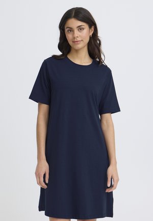 Ženska s temnimi lasmi, ki nosi ohlapno, kratkoročno mornarsko modro obleko, stoji na enostavni svetli podlagi.