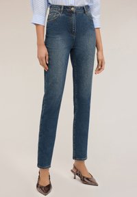 Niebieskie dżinsy z wysokim stanem o dopasowanym kroju, z przednimi kieszeniami i ozdobnym guzikiem. W połączeniu z błyszczącymi butami na obcasie.