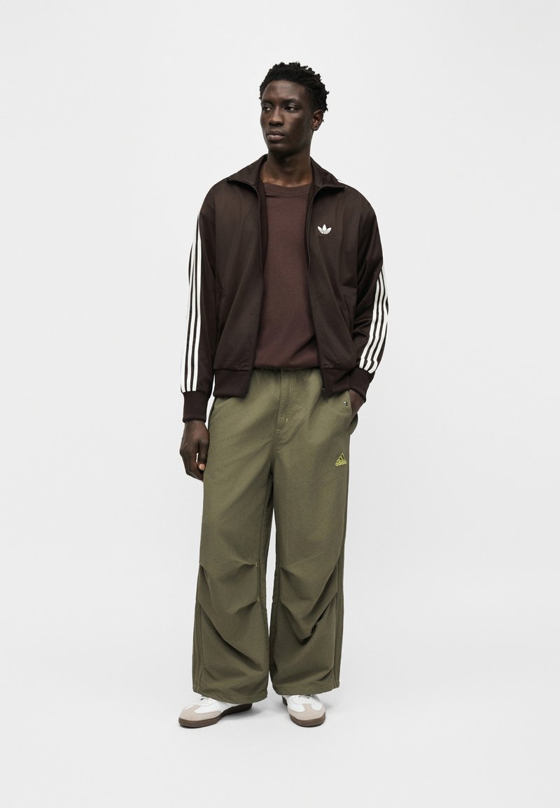 Uomo in piedi che indossa una giacca da track Adidas marrone scuro, una maglietta marrone, pantaloni larghi verde oliva con logo Adidas e scarpe da ginnastica bianche.