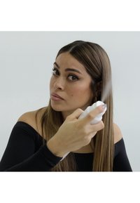 White spray bottle held in hand, releasing mist, with a model in a black off-shoulder top and long brown hair, against a light background.