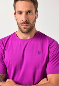 Krátký rukáv tričko v jasně fialové barvě; hladký materiál s kulatým výstřihem. Malé černé logo na levé hrudi. Bez vzorů nebo akcentů.
