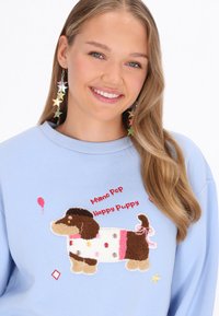 Jasnoniebieska bluza z teksturowanym, haftowanym jamnikiem w kropki i zabawnym napisem, uzupełniona kolczykami w kształcie gwiazdek.