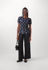 Blouse bleu marine à pois avec une taille peplum, manches courtes ballon ; associée à un pantalon gris à large jambe et des ballerines noires, portant un petit sac à main.
