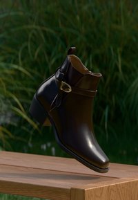 Glänzender, dunkelbrauner Knöchelstiefel mit seitlichem Schnallenverschluss und niedrigem Absatz, der auf einer Holzoberfläche liegt, mit unscharfem grünem Gras im Hintergrund.