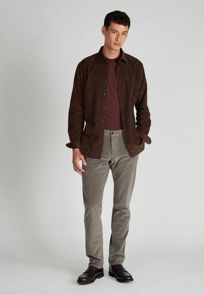 Camicia di velluto a coste marrone sopra una maglietta color bordeaux, abbinata a pantaloni grigi chiari con texture e mocassini scuri, in piedi davanti a uno sfondo chiaro.