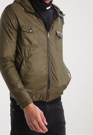 Homme portant une veste zippée vert olive avec poches poitrine et latérales, associée à une chemise noire et un pantalon noir.