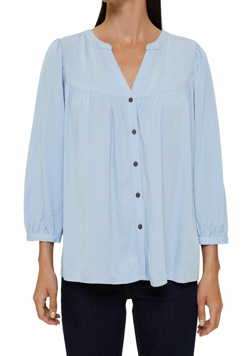 Blouse bleu clair avec un col en V, des détails de boutons et des manches bouffantes trois quarts. Fabriquée dans un tissu texturé avec une coupe ample.