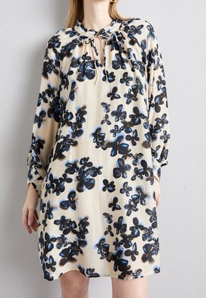 Kvinde iført en løs beige kjole med lange ærmer, sort og blå blomsterprint, og et lille forreste bånd ved halsudskæringen.