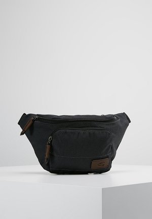 Sac banane en tissu noir avec deux compartiments zippés et tirettes marron, placé sur une surface blanche devant un fond gris uni.