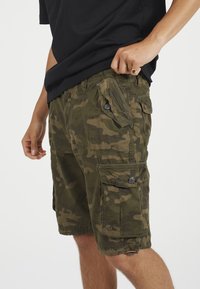 Persona che indossa pantaloni cargo mimetici verdi con molte tasche a bottoni e una maglietta nera, mentre sistema l'orlo della maglietta.