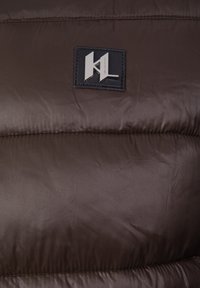 Sección de chaqueta acolchada marrón con una textura suave y brillante. Presenta una etiqueta rectangular negra con detalles de logotipo en plata.