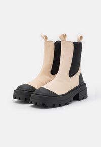 Botas Chelsea chunky com parte superior bege, painéis elásticos pretos, bico arredondado e sola de borracha preta texturizada com cravos profundos.