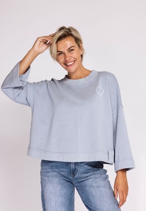 Lächelnde Frau mit kurzen blonden Haaren trägt einen lockeren hellgrauen Sweatshirt und blaue Jeans, sie berührt mit einer Hand ihr Haar.