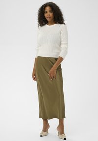 Femme aux cheveux bouclés portant un pull en tricot blanc, une jupe midi olive et des mules beiges à talons, se tenant sur un fond blanc.