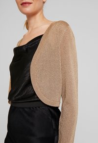 Guldig skimrande bolerosweater med långa ärmar, öppen framdesign och mjuk textur, bärs över en svart satintopp.