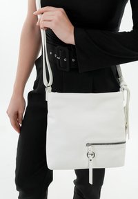 Bolso bandolera de cuero blanco con un bolsillo delantero con cremallera, forma rectangular y correa ajustable. Diseño simple con líneas suaves.