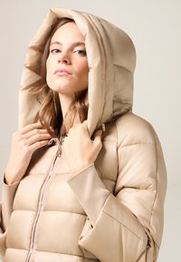 Puchowa beżowa kurtka z dużym kapturem, z zamkiem błyskawicznym na środku, ściągaczami na rękawach i przeszyciami pikowanymi dla izolacji.