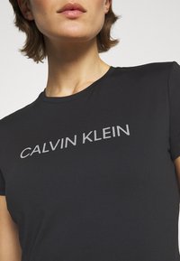 Camiseta negra de manga corta con cuello redondo. Presenta un logo plateado de "CALVIN KLEIN" en el pecho. Textura de tela suave.