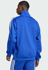 Blaues Zip-Jackett aus glattem Stoff, mit hohem Kragen und weißen Streifen an den Ärmeln. Lässige Passform mit minimalen Details.