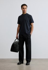 Σκούρο μπλε μπλουζάκι με ψηλό λαιμόκοψη και διακριτική σήμανση στον γιακά, σε συνδυασμό με φαρδιά μαύρη παντελόνια. Το μοντέλο κρατά μια μαύρη δερμάτινη τσάντα.