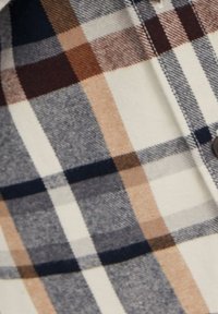 Camicia a scacchi caratterizzata da motivi a quadri blu, marroni e beige su uno sfondo crema. Realizzata in tessuto morbido con dettagli con bottoni.