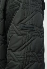 Μαύρο καπιτονέ ύφασμα με ραφές ζιγκ-ζαγκ και μικρό ορθογώνιο ταμπελάκι με την ένδειξη "KARL LAGERFELD" σε μανίκι ρούχου.