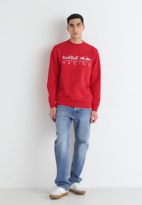 Sudadera roja con el logo de "Red Bull Racing", combinada con jeans de color azul claro. Zapatillas blancas con suelas marrones completan el conjunto.
