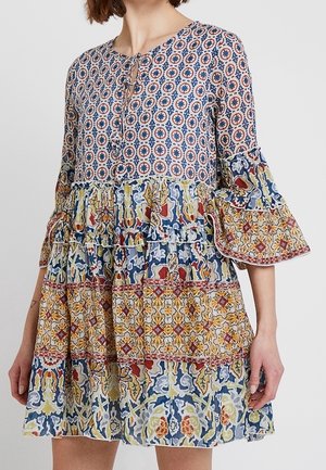 Kobieta w wielokolorowej, boho sukience z okrągłymi i kwiatowymi wzorami, rękawami o długości 3/4 z falbankami oraz krótką, warstwową spódnicą.