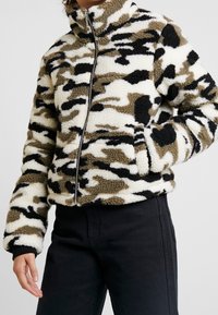 Luddig, zip-up jacka i ett kamouflagedesign med svarta, beiga och vita mönster. Har hög krage och sidofickor. Accentuerad med ribbade ärmslut.