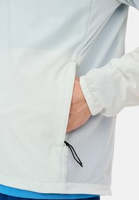 Giacca leggera e traspirante in blu pallido e bianco, con tessuto in rete, polsini elastici e tasche laterali con cordoncino nero.