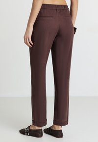 Pantaloni marroni leggeri e tessuti, con vestibilità rilassata, vita elastica e orli arrotolati, dotati di due tasche posteriori. Abbinati a scarpe nere.