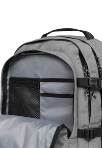 Mochila de tela gris con un bolsillo de malla y múltiples compartimentos, con correas y cremalleras negras, que presenta un exterior texturizado.