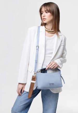 Joven mujer con blazer blanco y jeans sostiene un bolso azul claro con correa de cuero, lleva un collar grueso de oro sobre un fondo liso.