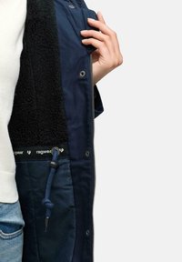 Navy jacket featuring a fuzzy black interior, zip closure, inner drawstring, and branded accents. Detail of hand holding the jacket.