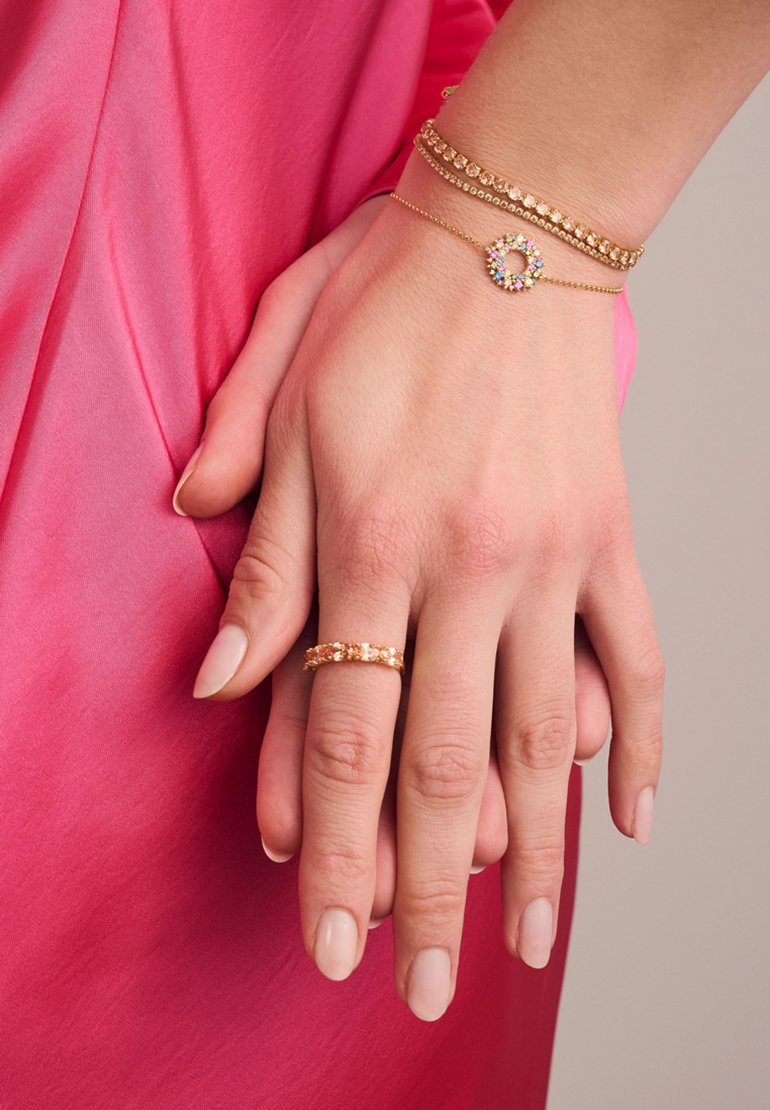 Des bracelets en or et une bague assortie avec des accents colorés sur une main, sur un fond en tissu rose lisse.