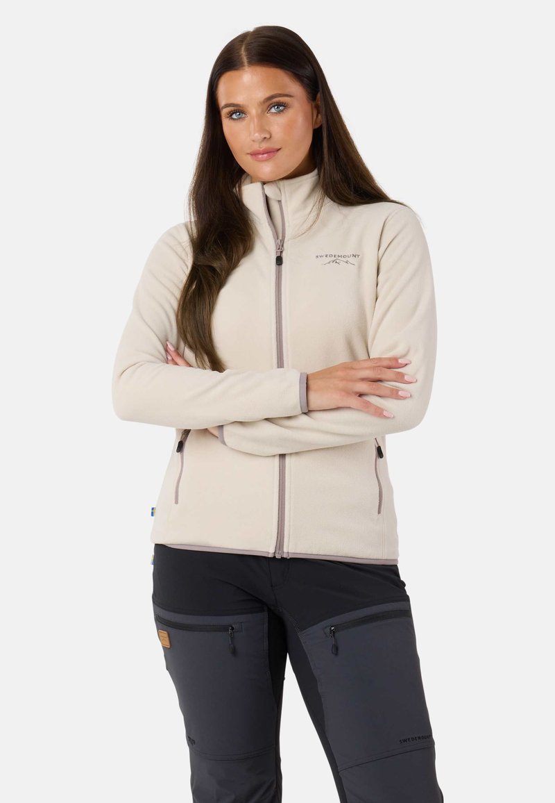 Vaalea beige fleece-takki, jossa on korkea kaulus ja vetoketjusulku, yhdistettynä mustiin housuihin, joissa on sivuvetoketjut ja logo-aksentti.